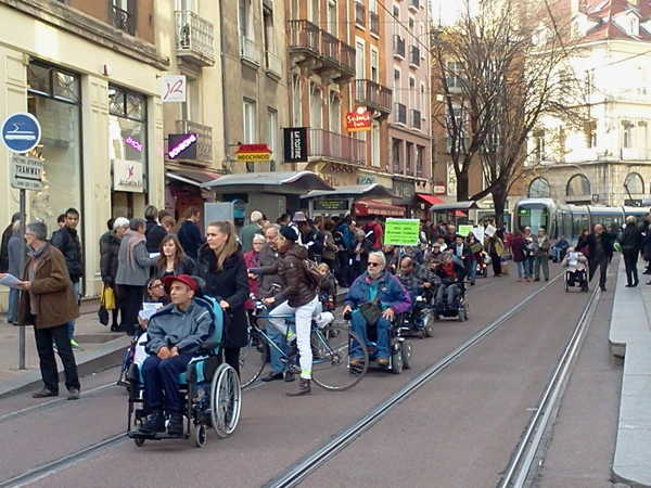 Sur cette photo, on voit des personnes handicapées en fauteuil roulant et d’autres personnes manifester en passant sur les rails du tramway au centre-ville. Des manifestants distribuent des tracts aux usagers, nombreux à la station de tram. On voit quelques pancartes.