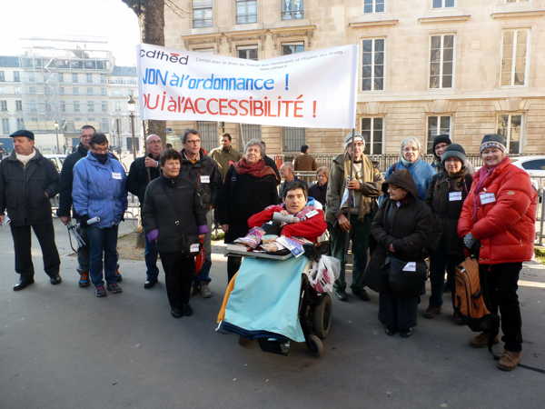 On voit une vingtaine de personnes chaudement vêtues, dont une en fauteuil roulant, alignées devant une banderole avec les inscriptions « CDTHED – non à l'ordonnance, oui à l'accessibilité ! » En arrière plain, deux immeubles haussmanniens.